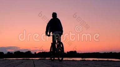 骑自行车的剪影。 少年骑自行车沿着河堤前行.. 体育生活方式。